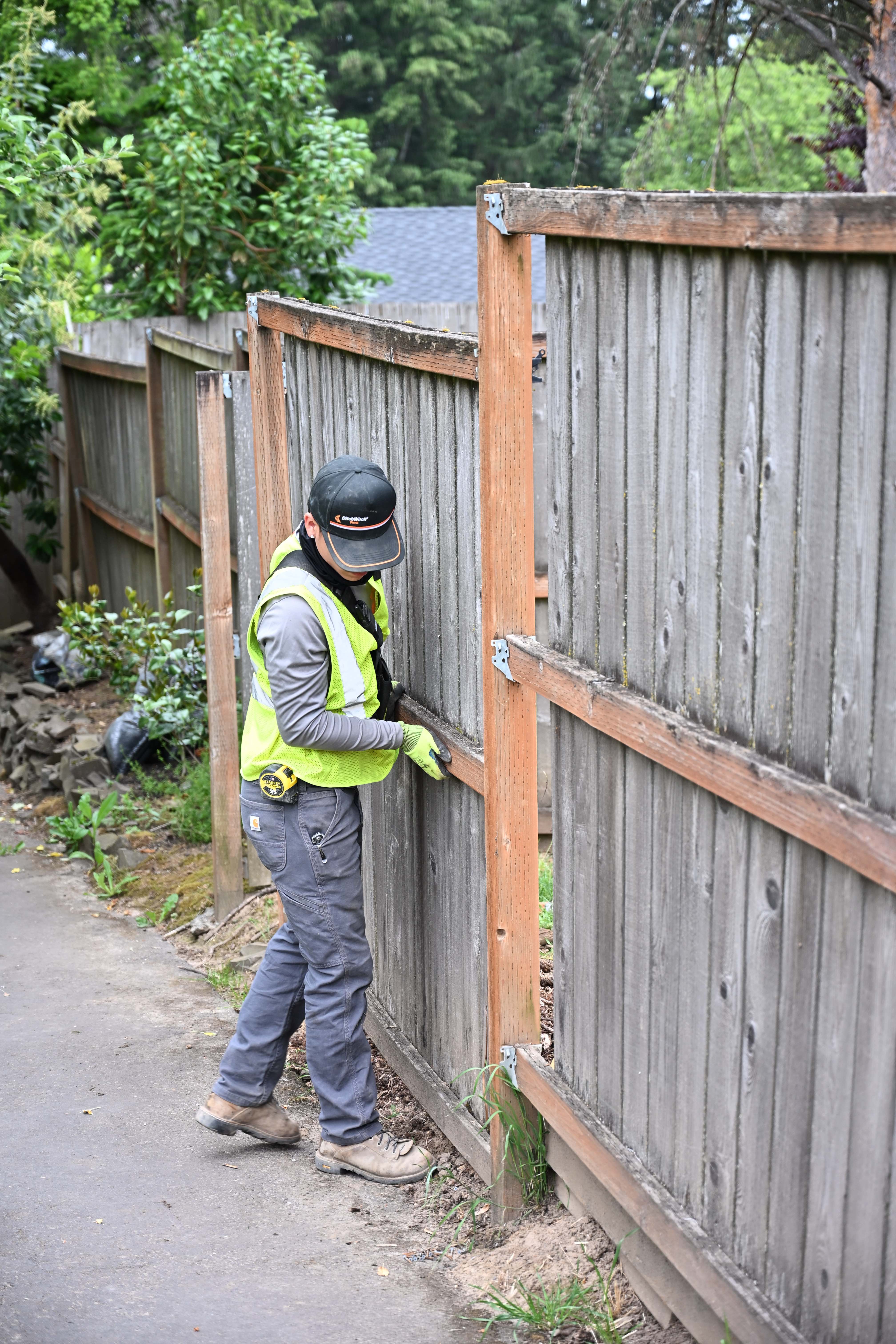 OCL crew member fitting fence rails and bracket hardware on a cedar fence line along a paved path