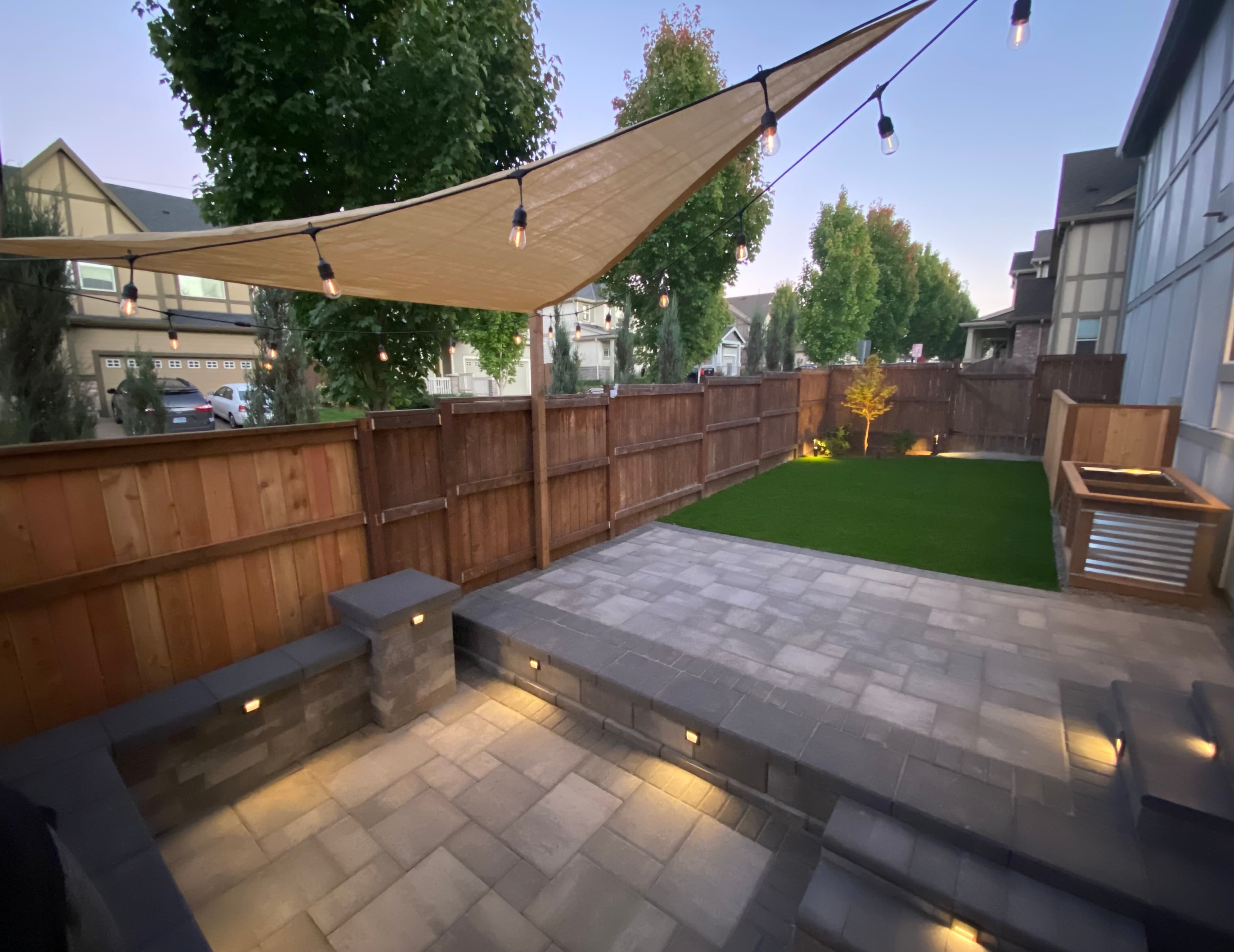 Finished multi-level OCL paver patio at dusk with integrated step lighting, seat wall, shade sail, and turf lawn