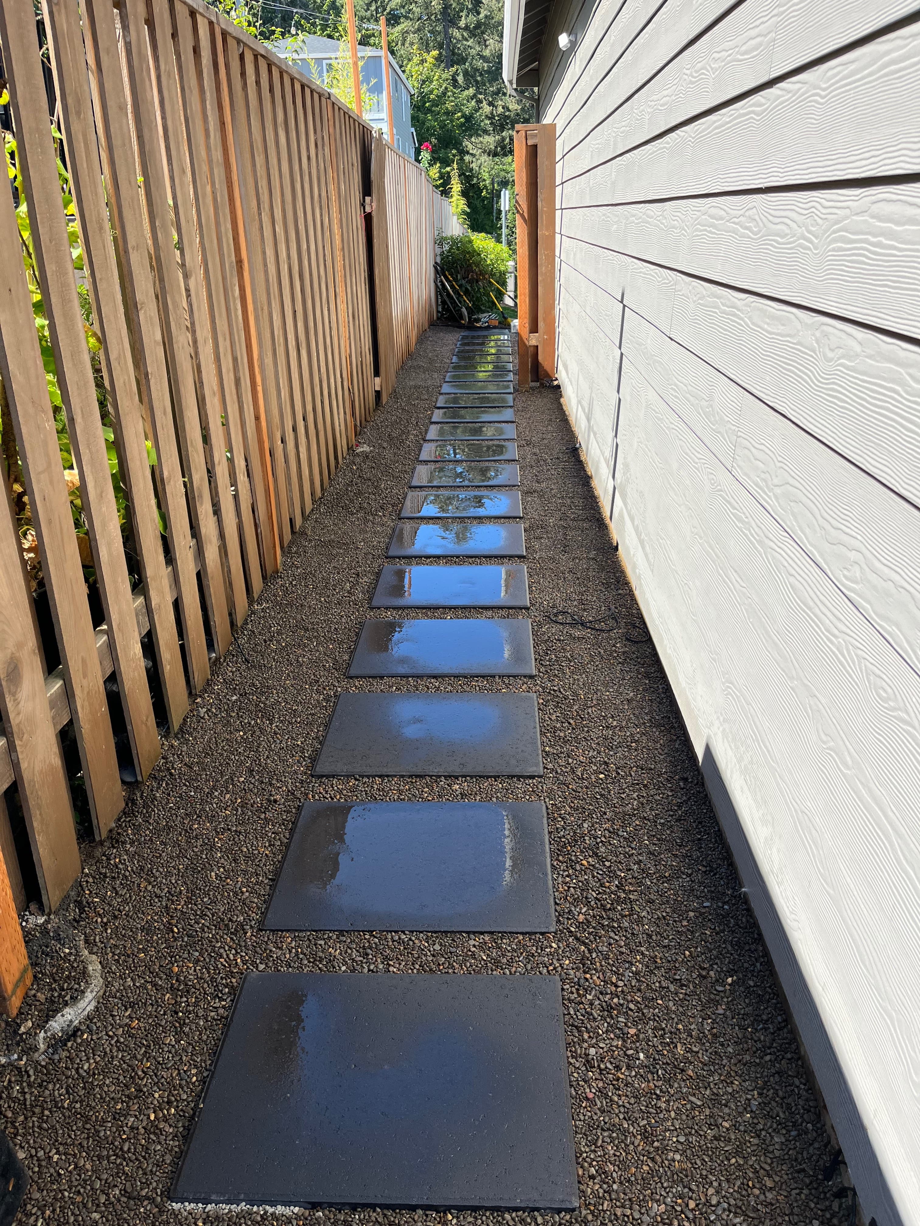 Side-yard stepping-stone path of large dark square pavers set in a gravel bed, running between a wood fence and home siding