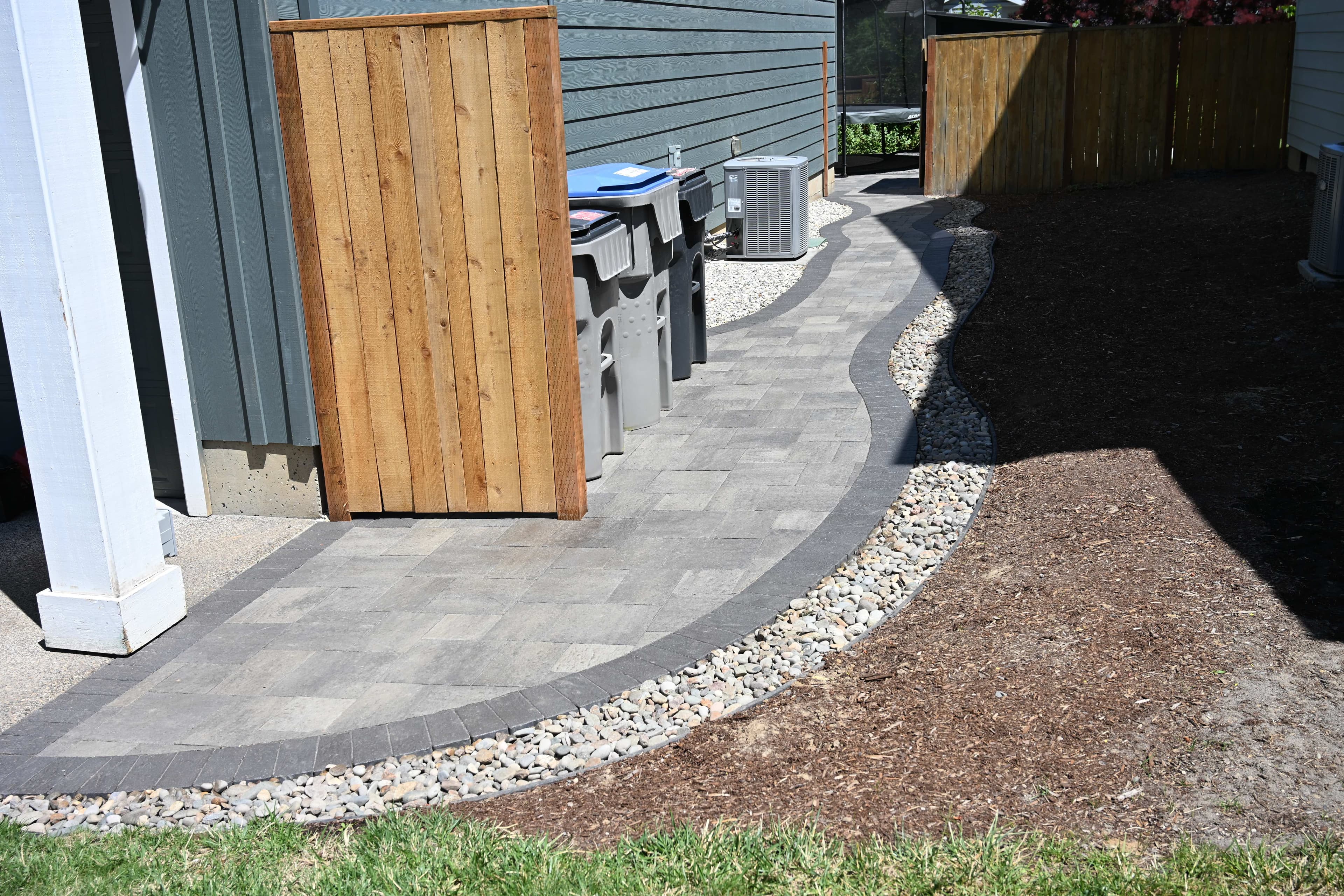 Curved paver walkway with a charcoal soldier-course border running alongside a home, framed by a river-rock drainage strip