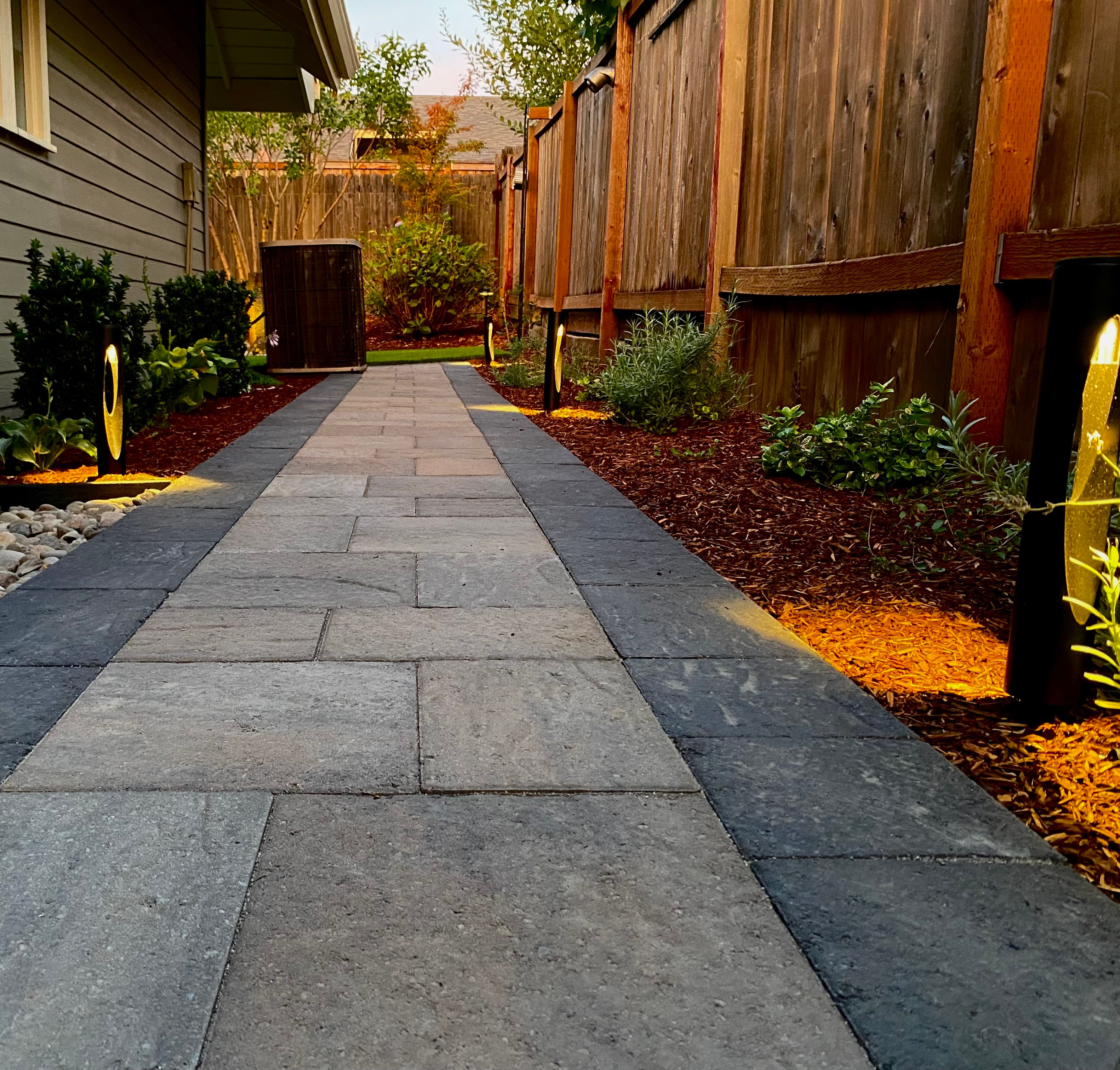 Dusk paver walkway with a charcoal soldier-course border, glowing path lighting, and mulched planting beds along a privacy fence