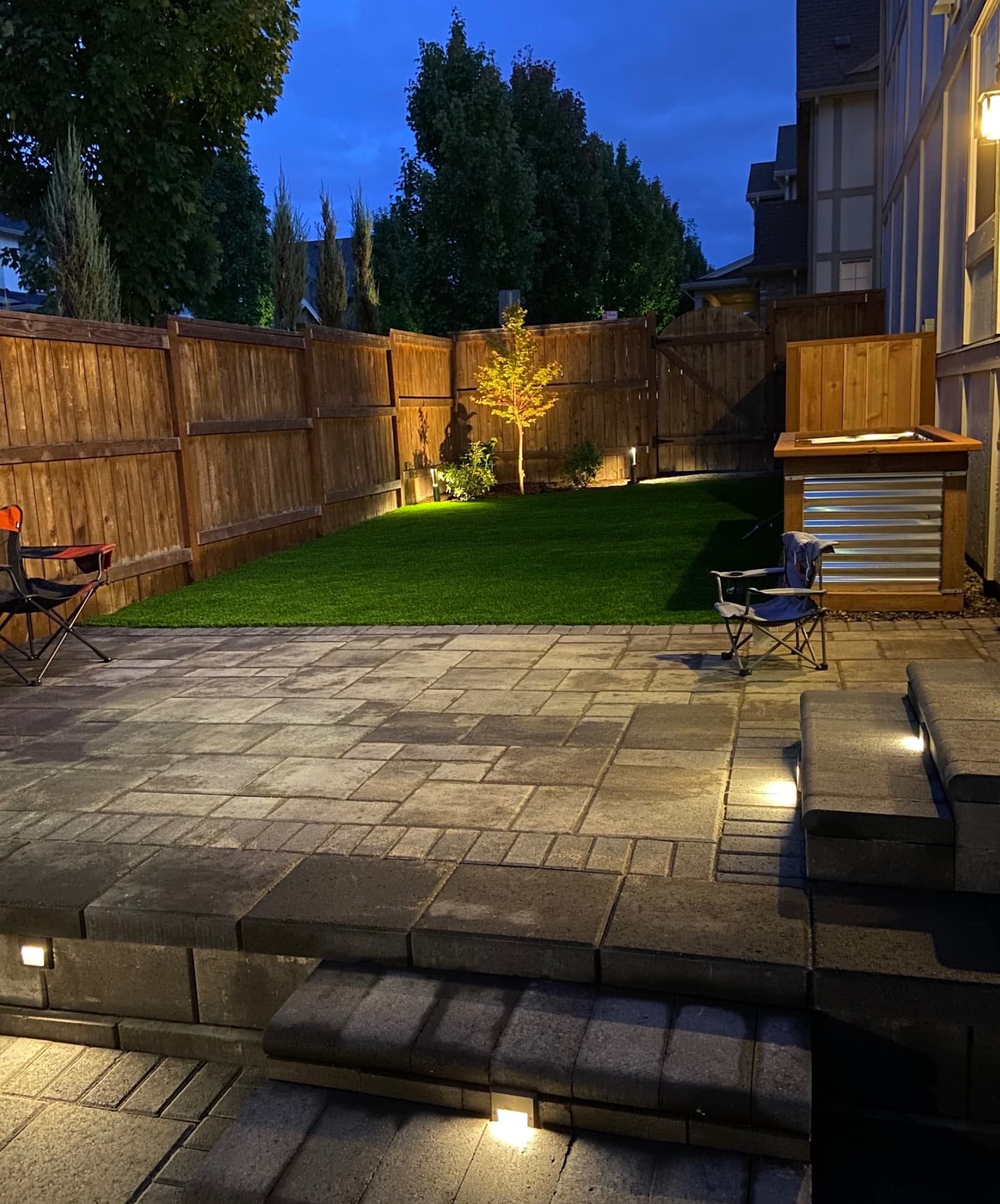 Backyard living area at dusk with a multi-level paver patio, step lighting, turf lawn, and an uplit ornamental tree