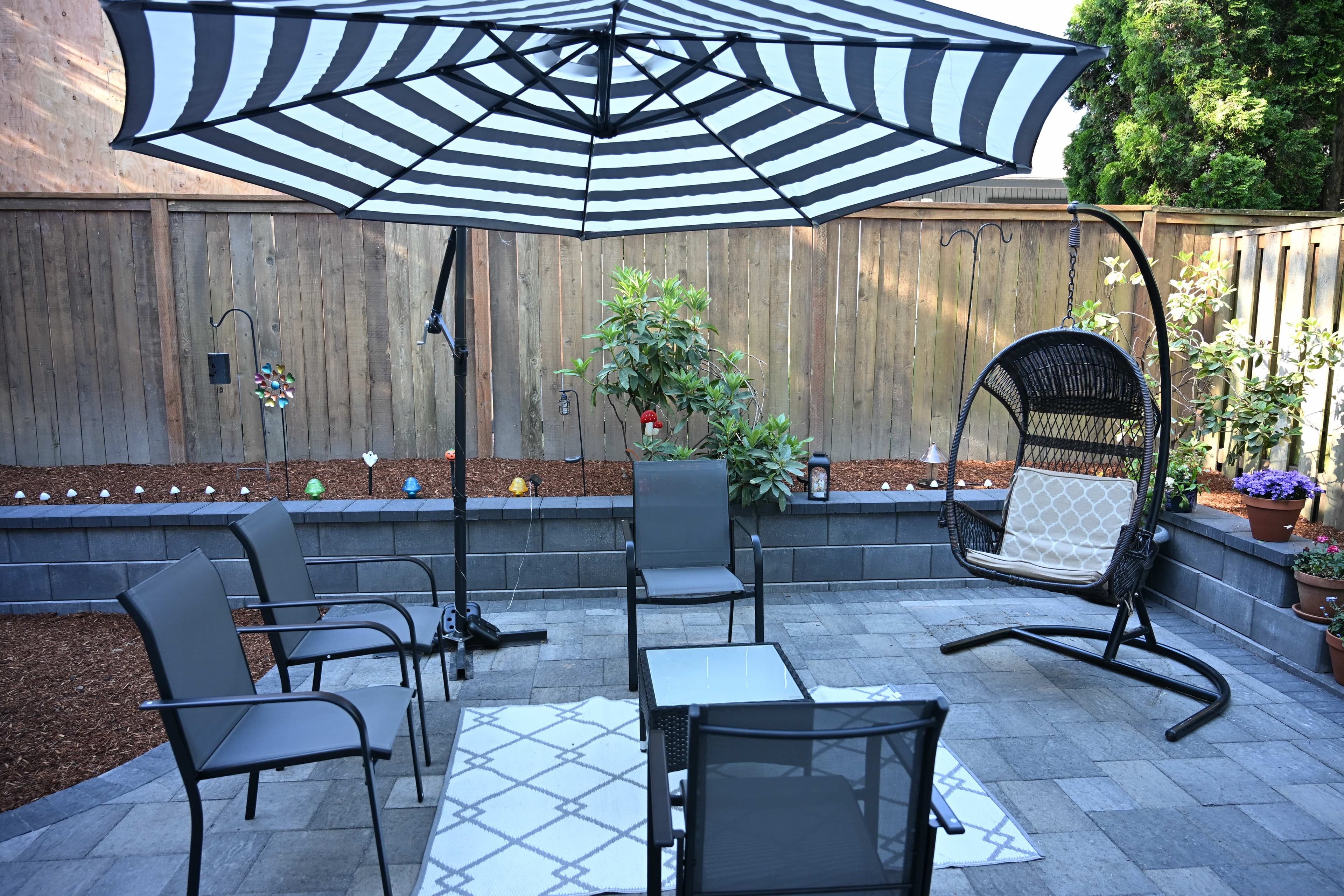 Finished backyard paver patio living area with a striped umbrella, mesh chairs, hanging egg chair, and block retaining wall