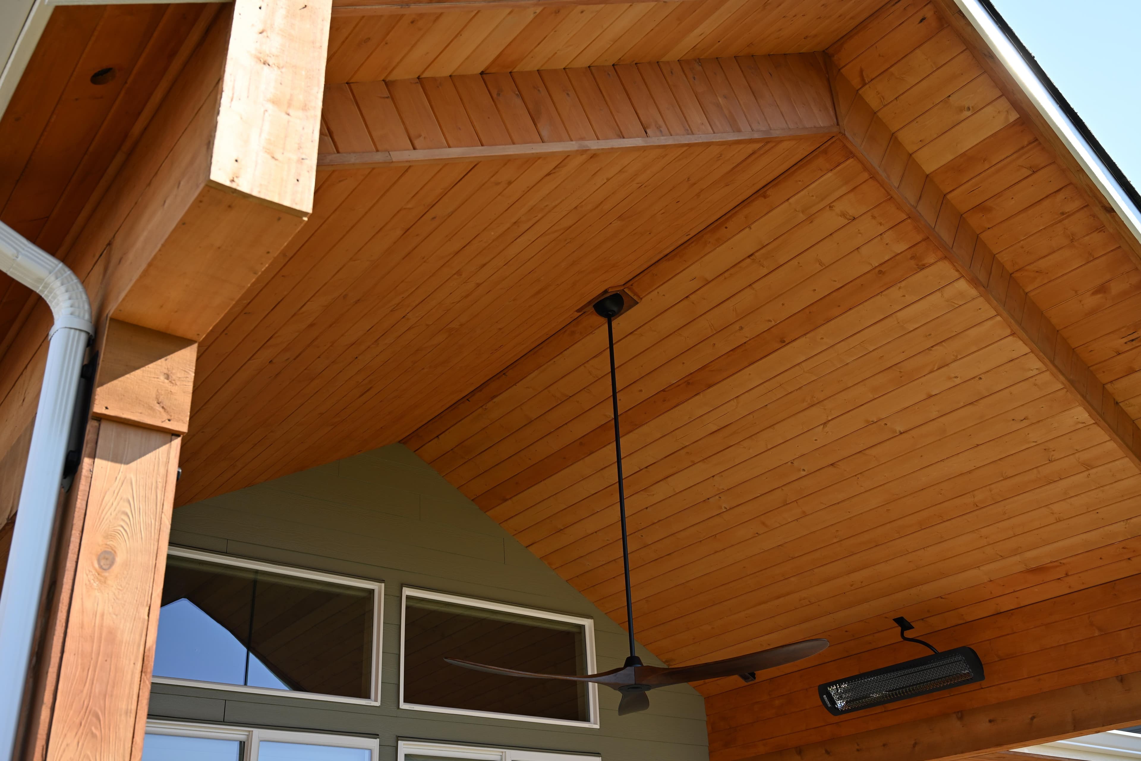 Upward view of a cedar tongue-and-groove gable patio cover ceiling with exposed beams, a black ceiling fan, and a patio heater on a sage-green home