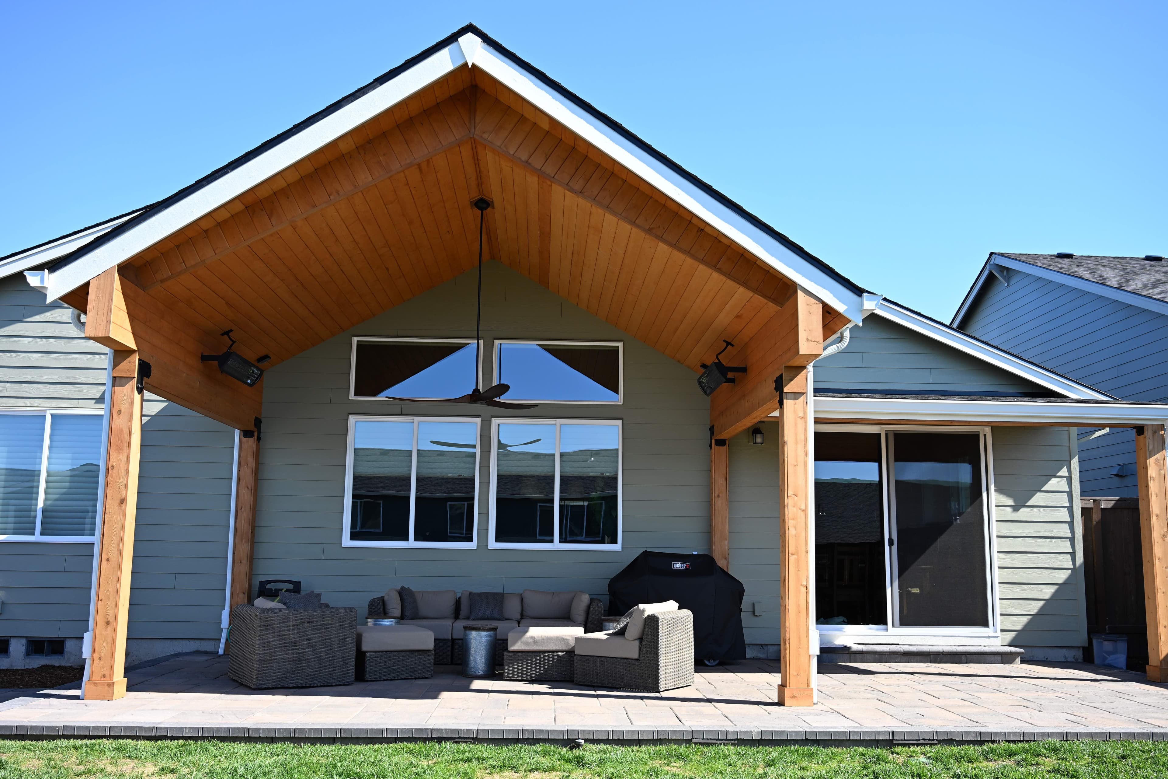 Cedar gable-roof pergola attached to a sage-green home with exposed wood ceiling, ceiling fan, and cedar posts over a paver patio