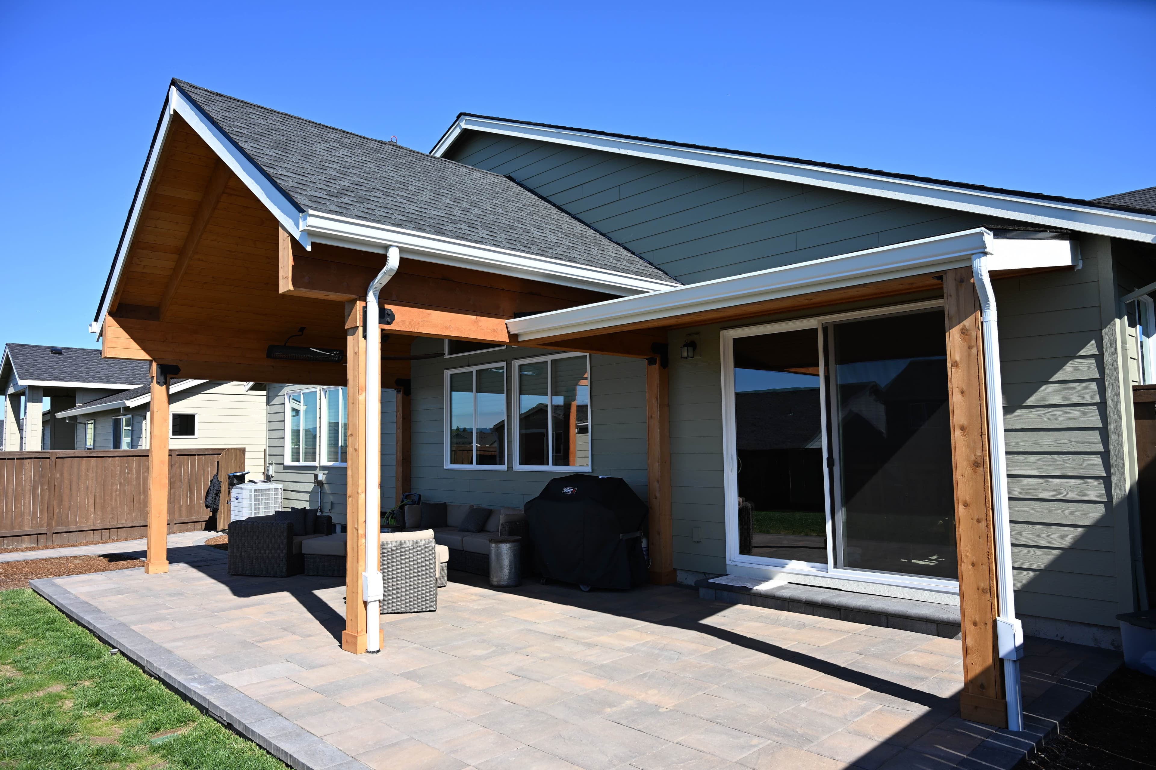 Cedar-post gable patio cover attached above a sliding-door entrance over a paver patio with outdoor seating and a grill