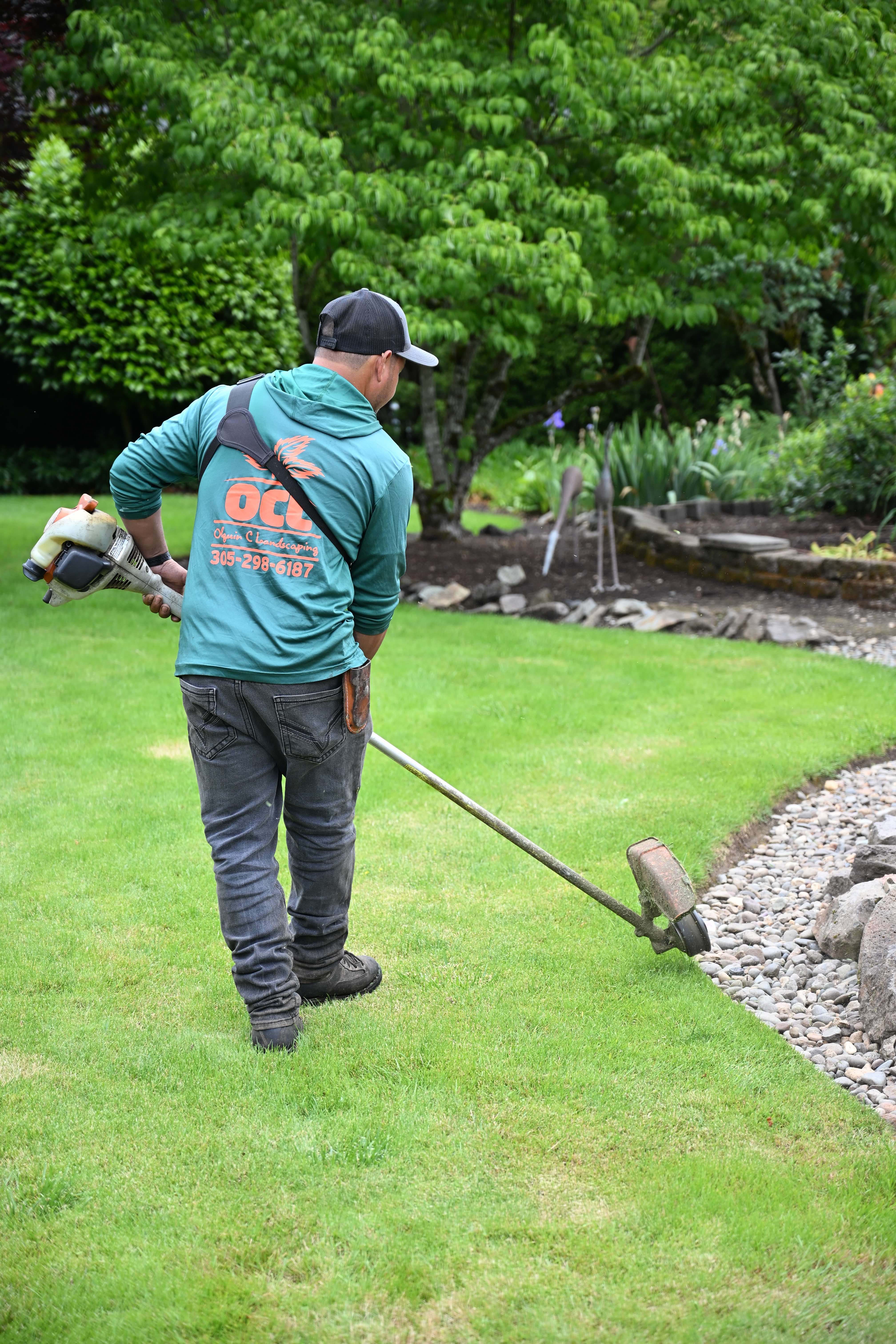 OCL crew member in branded teal hoodie edging a vibrant green lawn along a river-rock border with a string trimmer