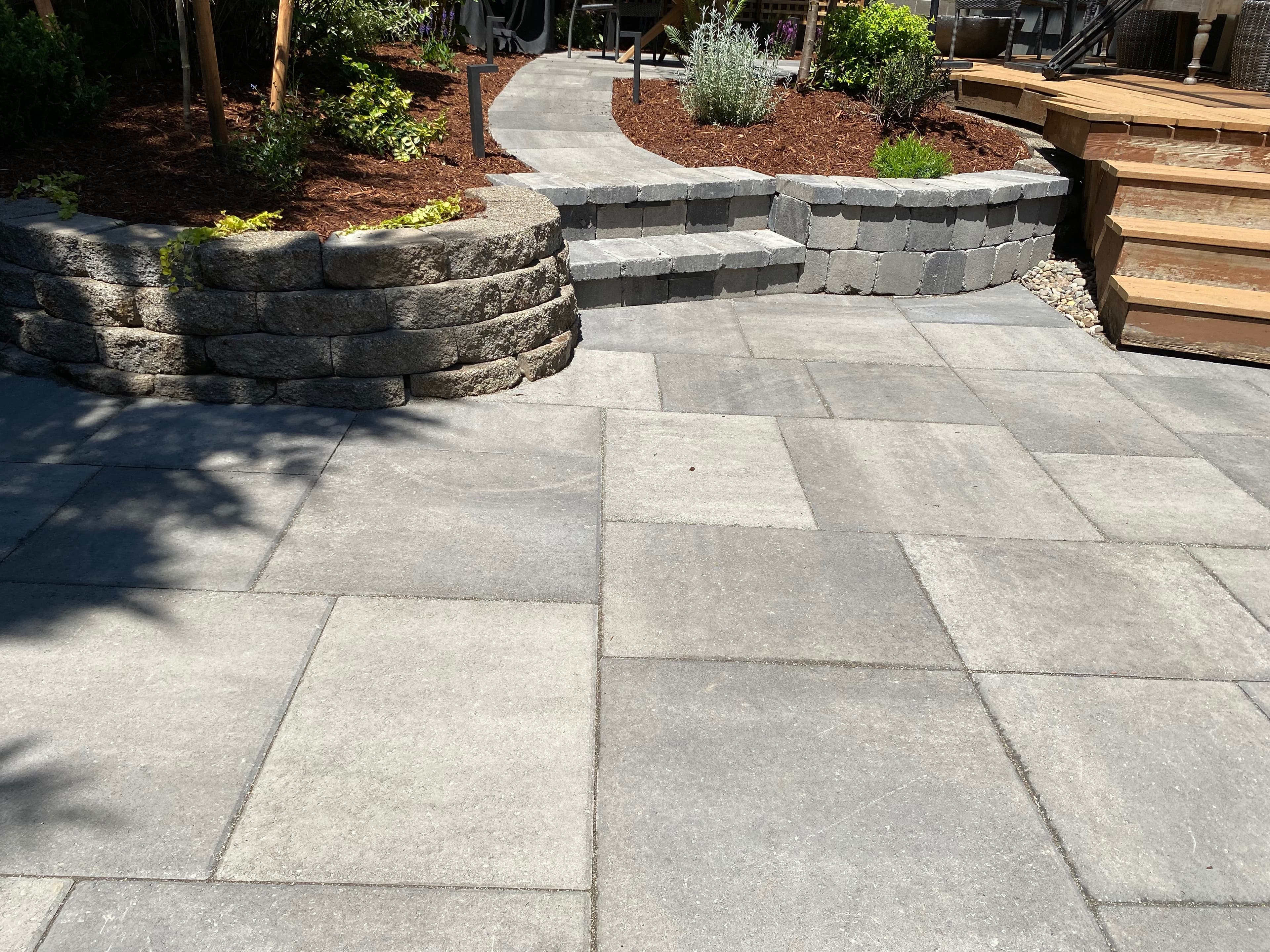 Curved gray segmental-block retaining and seat walls with integrated steps framing raised planting beds and a large-format paver patio