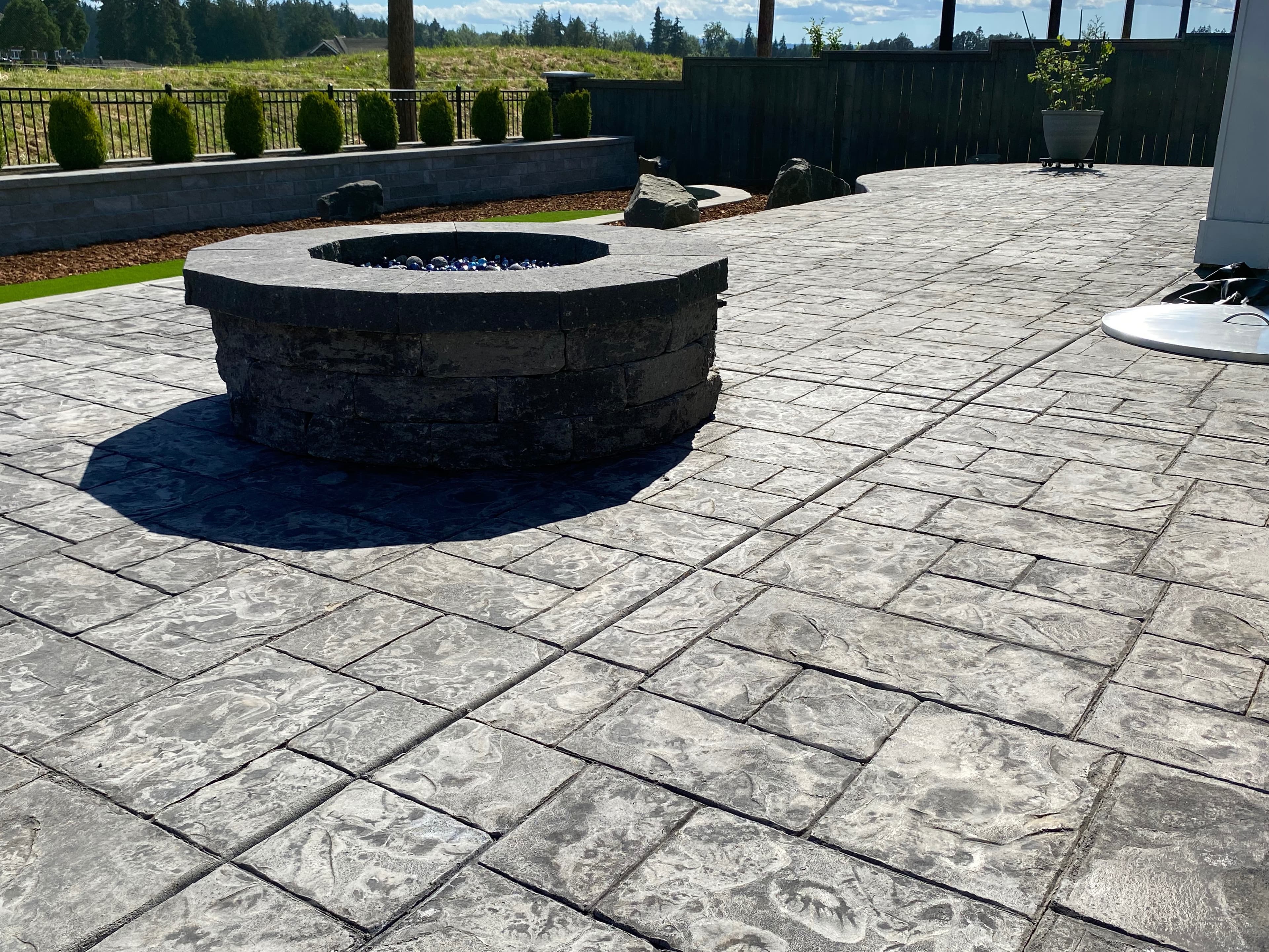 Stacked-stone gas fire pit with a low block seat wall on a stamped-concrete patio overlooking an open field