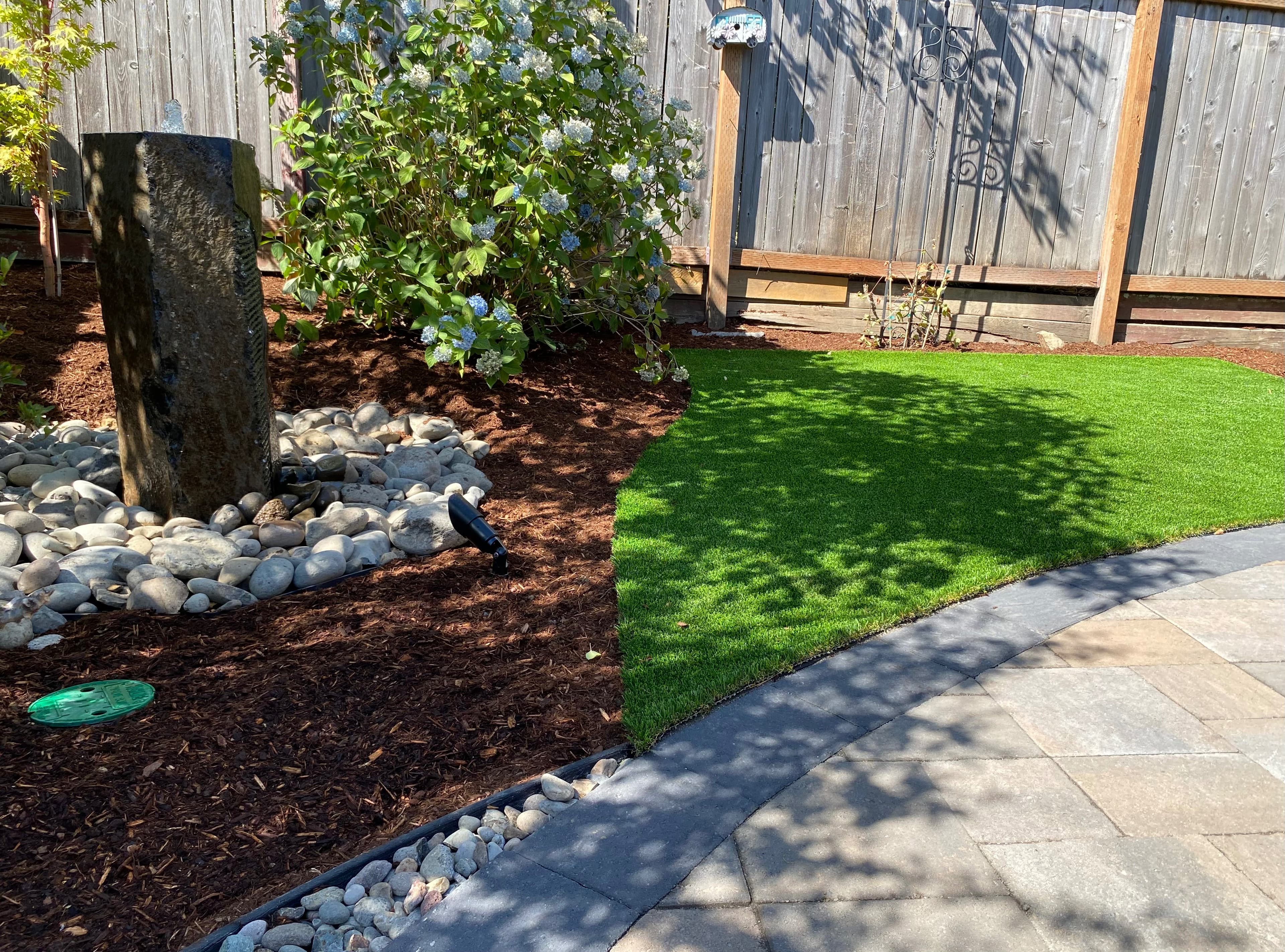 Tall basalt column fountain set in a river-rock bed beside a blue hydrangea, with a manicured lawn and paver path